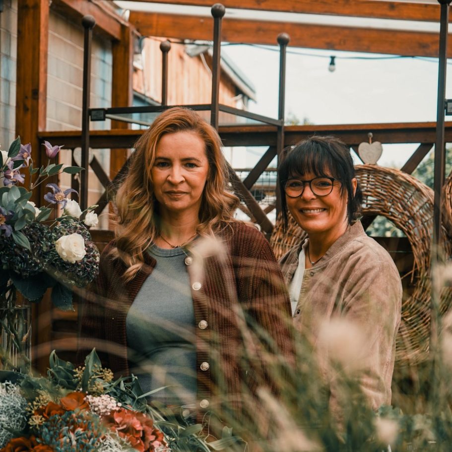 blumenundideen_teamfoto Zwei Frauen stehen entspannt inmitten von Blumen und Pflanzen auf einem romantischen Hintergrund.