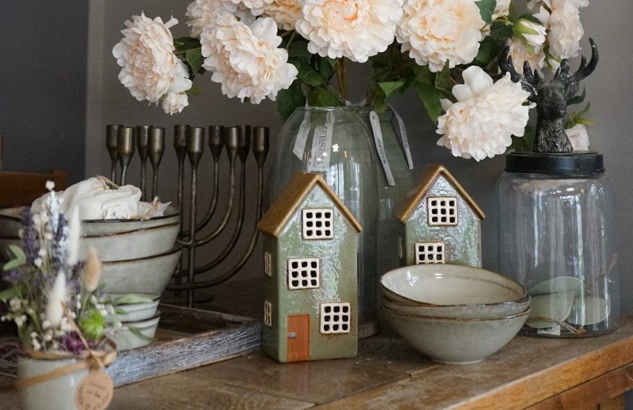 blumenundideen_wohnaccessoires Zwei kleine Keramikhäuser, Blumen in Vasen und Schalen auf einem Holztisch.