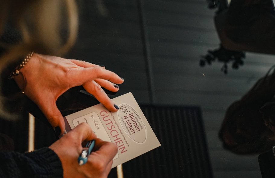 blumenundideen_Gutschein Eine Hand schreibt auf eine Gutscheinkarte, umgeben von einem Holzboden und Pflanzen.